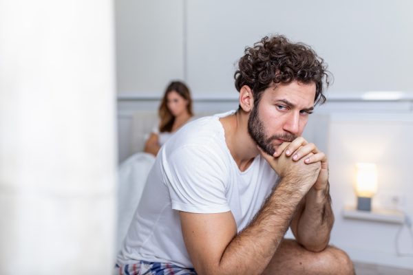 Sad thoughtful man after arguing with girlfriend.Relationship difficulties, conflict and family concept,unhappy couple having problems at bedroom. Sad guy sitting on bed,girlfriend in the background