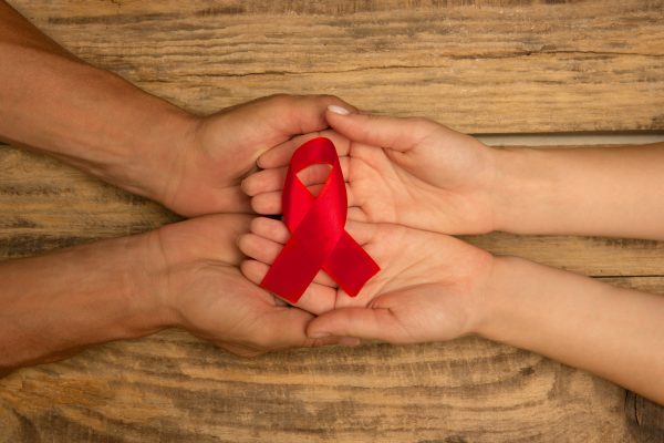 Weibliche und männliche Hände halten rotes HIV- und AIDS-Sensibilisierungsband isoliert auf Holzhintergrund. Konzept der Gesundheitsversorgung und Medizin, weltweit unterstützende, ehrenamtliche Kampagne gegen Krankheiten.